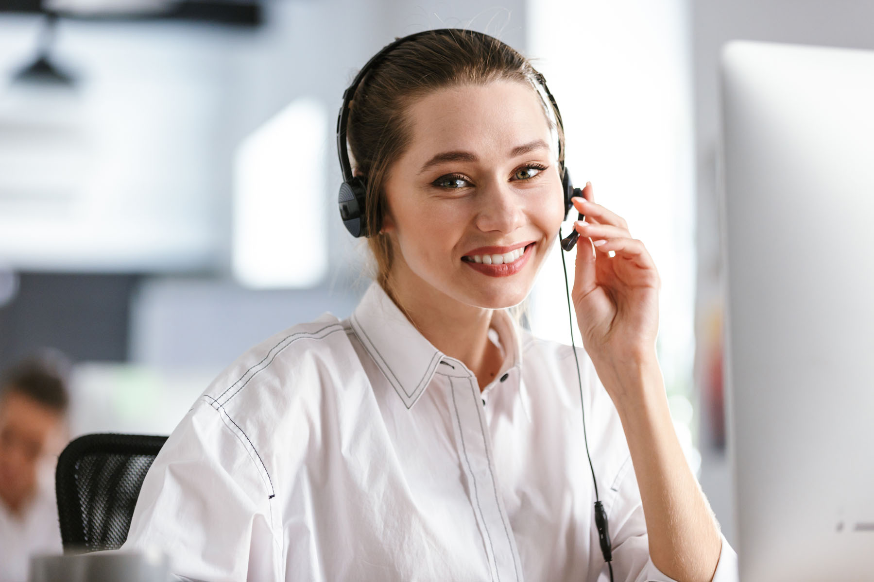 Happy young woman wearing microphone headset dressed in shirt sitting at her workplace at the office, speaking with client